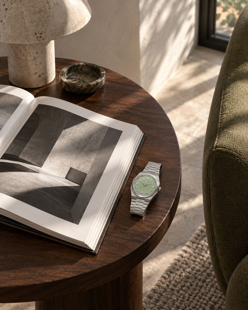 Vale 03 Steel / Green beside an open art book on a round side table in a quiet reading corner